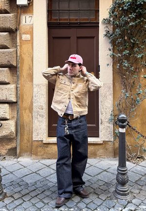 Hombre con chaqueta beige, gorra rosa y vaqueros oscuros de pie en una calle empedrada frente a una puerta marrón y una pared amarilla con plantas.