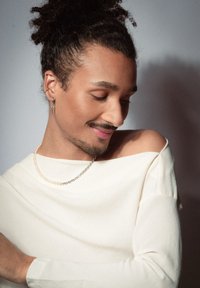 Witte, off-shoulder top met een zachte textuur; gelaagde sieraden met een parel en zilveren ketting; haar is gestyled in een krullende updo.