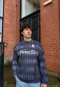 Long-sleeve shirt in shades of grey with a diamond pattern, featuring the "Fantazy FC" logo and a round neckline. Worn with light blue jeans.