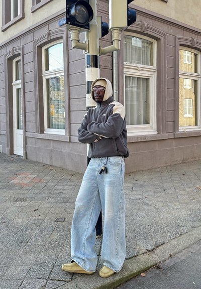Chaqueta de forro polar gris y beige, vaqueros de pierna ancha azul claro y zapatos slip-on beige. De pie junto a un semáforo frente a un edificio marrón.