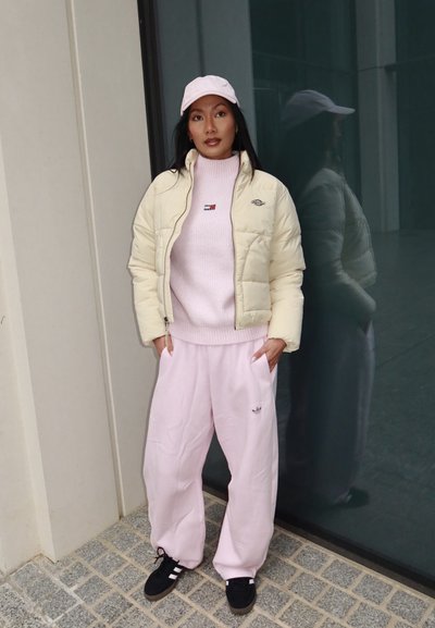 Mujer con chaqueta acolchada color crema, suéter rosa, pantalones rosas y gorra rosa, de pie con las manos en los bolsillos frente a una pared de vidrio que refleja su imagen.