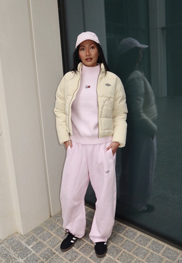 Naine, kes kannab kreemjat sulejope, roosat kampsunit, roosasid pükse ja roosat mütsi, seisab käed taskutes klaasseina ees, mis peegeldab tema kuju.