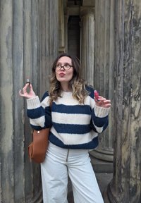 Strickpullover mit blauen und weißen Streifen, beigefarbene weite Hose, hält ein rosa Lippenprodukt. Der Hintergrund zeigt Steinsäulen.
