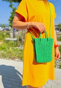 Bolsa tecida verde feita de material texturizado, com duas alças curtas e uma alça longa, combinada com um vestido laranja simples.