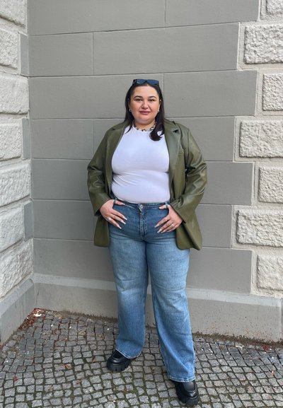 Blazer de cuero verde oliva sobre una camisa blanca, combinado con unos vaqueros de pierna ancha en azul claro y unos zapatos negros gruesos. Detalles de joyería pequeña en oro visibles.