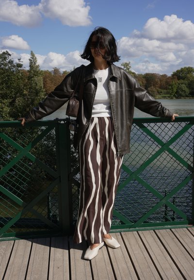 Chaqueta de cuero marrón sobre una blusa blanca, combinada con pantalones de rayas anchas en marrón y blanco. Zapatos planos blancos y bolso pequeño. Tarima de madera y barandilla verde.