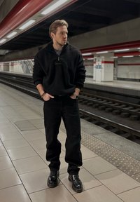 Juoda pūkinė pusiau užtraukiama striukė, juodos kelnės ir juodi batai. Lygi tekstūra, laisvas siluetas. Metro platforma su raudonais ir pilkais elementais fone.