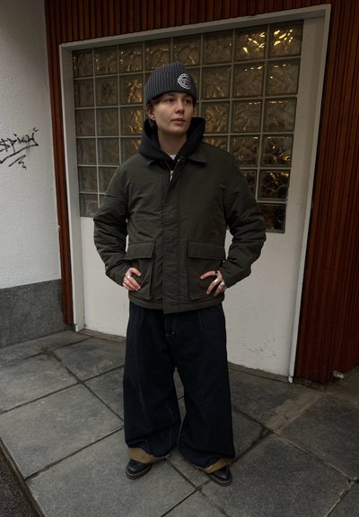 Persona con chaqueta verde oscuro, sudadera con capucha negra, gorro gris, pantalones anchos oscuros y zapatos negros, de pie con las manos en las caderas frente a un edificio.