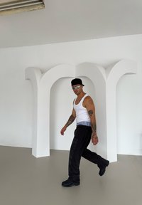 Calças pretas com um cós riscado, top branco em formato de tanque e botas pretas. A caminhar ao lado de uma estrutura de arco abstrata branca sobre um chão claro.