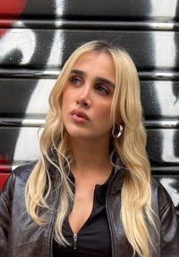 Eine Person mit langen blonden Haaren, die ein schwarzes Zip-Up-Oberteil und eine dunkle Lederjacke trägt, vor einem Graffiti-Hintergrund in Rot- und Schwarztönen.