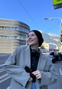 Blazer gris surdimensionné avec des manches larges, chemise noire en dessous, écouteurs noirs, tenant des lunettes de soleil, arrière-plan urbain avec des montagnes.