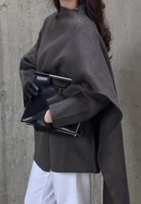 Grå ullponcho med høy hals, båret over hvite bukser. Svart clutch holdt i hanskekledde hender. Glatte teksturer og subtile designdetaljer.