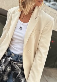 Kremowa marynarka z wykładanym kołnierzem, zestawiona z białym topem na ramiączkach z czarnym logo "HUGO" oraz dżinsowymi szortami, z kraciastą koszulą związaną w talii.