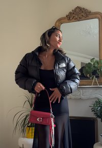 Donna che indossa un giubbotto imbottito nero North Face e un vestito nero trasparente, mentre tiene una borsa rossa, in piedi accanto a uno specchio con cornice dorata e piante.
