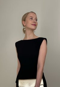 Vestido negro ajustado con mangas cortas tipo capa, textura lisa. Combinado con un bolso de mano metálico dorado y pendientes colgantes brillantes sobre un fondo neutro.