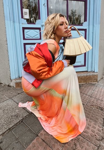 Mujer agachada al aire libre con un vestido degradado de colores y una chaqueta naranja, sosteniendo un bolso beige plisado cerca de una puerta azul y púrpura.