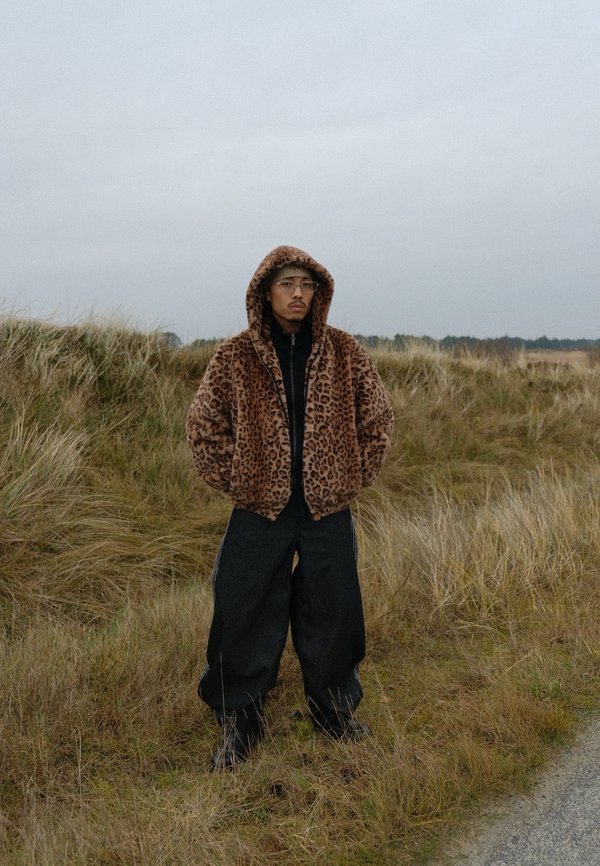Braune Faux-Felljacke mit Leopardenmuster, Reißverschluss vorne und Kapuze. Dazu schwarze, weit geschnittene Hosen mit seitlichen Streifen. Hintergrund mit Grasland.