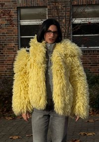 Gul jacka i konstpälss med lurvig textur, dragkedja och överdimensionerade ärmar. Bärs över en grå topp och kombineras med grå jeans.