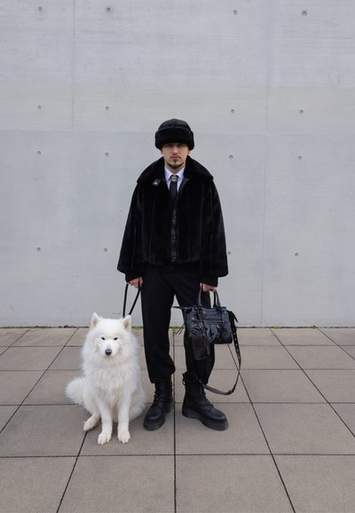 Chaqueta de piel negra con un cuello alto, pantalones negros, botas negras y un accesorio metálico. Un perro blanco y esponjoso está sentado a los pies de la persona.