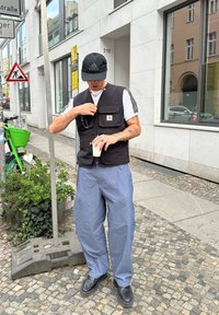 Črna utilitarna brezrokavnica z več žepi, oblačena čez belo prugasto srajco, v kombinaciji z ohlapnimi modrimi kavbojkami in črnimi čevlji. Zunanje urbano okolje.
