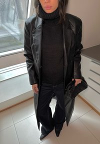 Černý kožený trench coat přes černý žebrovaný rolák, doplněný tmavými džíny a černými lodičkami s špičatou špičkou. Doplňkem je pletená černá kabelka.