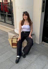 Μαύρα παντελόνια σε φαρδιά γραμμή, άσπρο τοπ χωρίς τιράντες, μαύροι loafers και μπεζ πλεκτή τσάντα με καφέ λεπτομέρειες, ακουμπισμένα σε μια κολόνα.