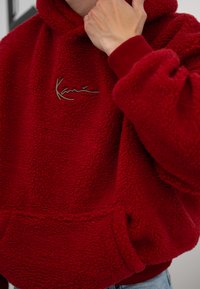 Κόκκινη φλις μπλούζα με κουκούλα, διαθέτοντας υφή, φαρδιά εφαρμογή, ριμπ μανσέτες και διακοσμητικό υπογεγραμμένο λογότυπο στο στήθος.