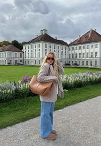 Ruskea nahkalaukku, jossa rento muotoilu; ruudullinen bleiseri valkoisen topin päällä; leveät siniset farkut; vaaleanruskeat kengät; taustalla kukkia ja historiallinen rakennus.