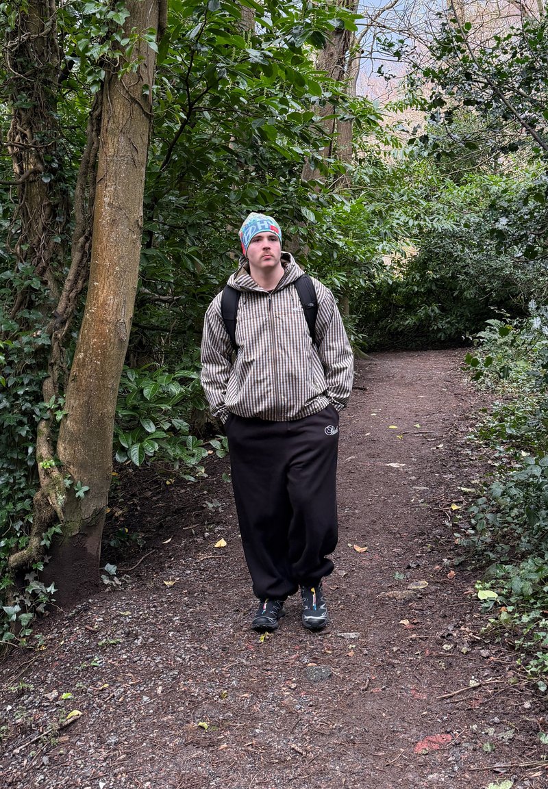 Hombre con chaqueta de cuadros, pantalones negros y gorro colorido caminando con las manos en los bolsillos por un sendero forestal rodeado de follaje verde.
