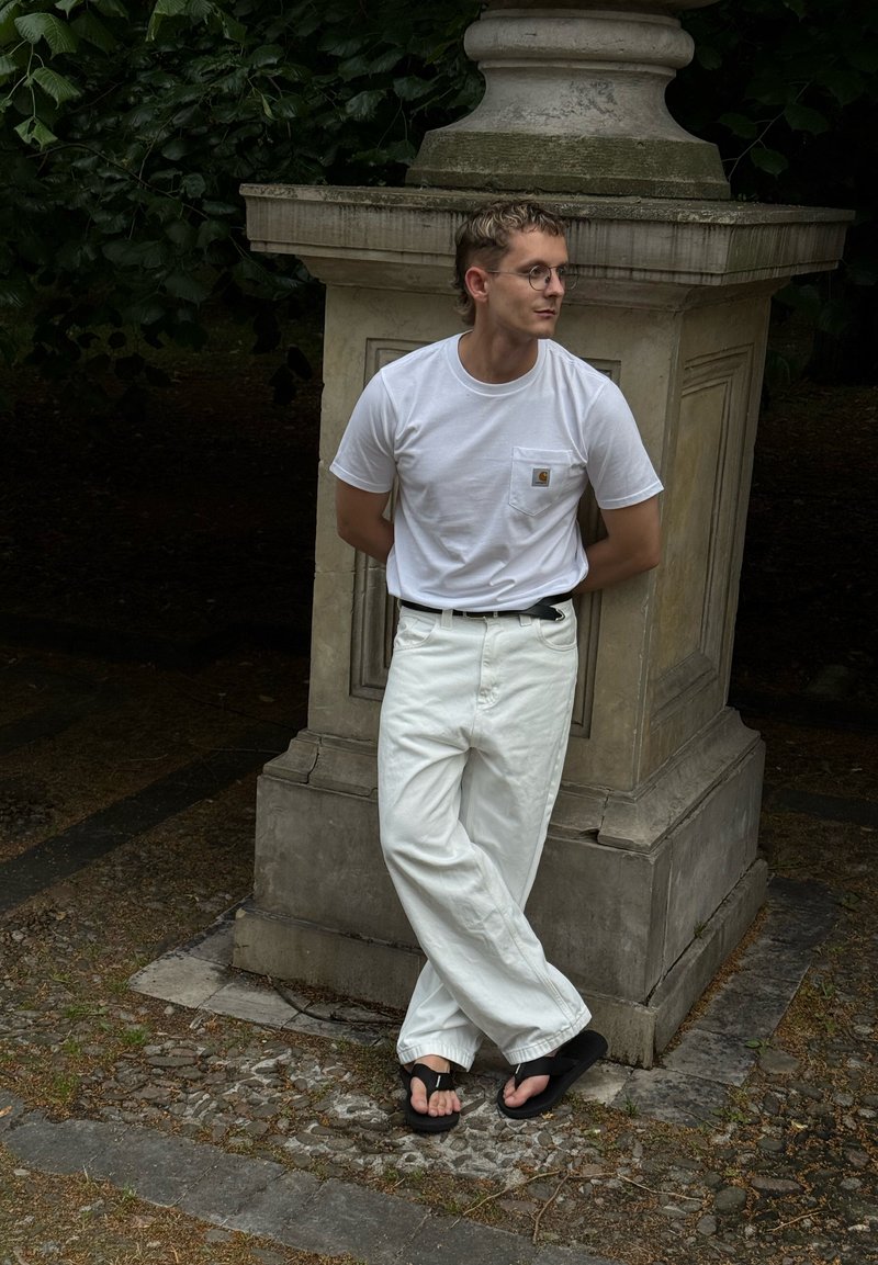 Jeune homme avec des lunettes, chemise blanche et pantalon, portant des tongs noires, appuyé contre un grand piédestal en pierre à l’extérieur.