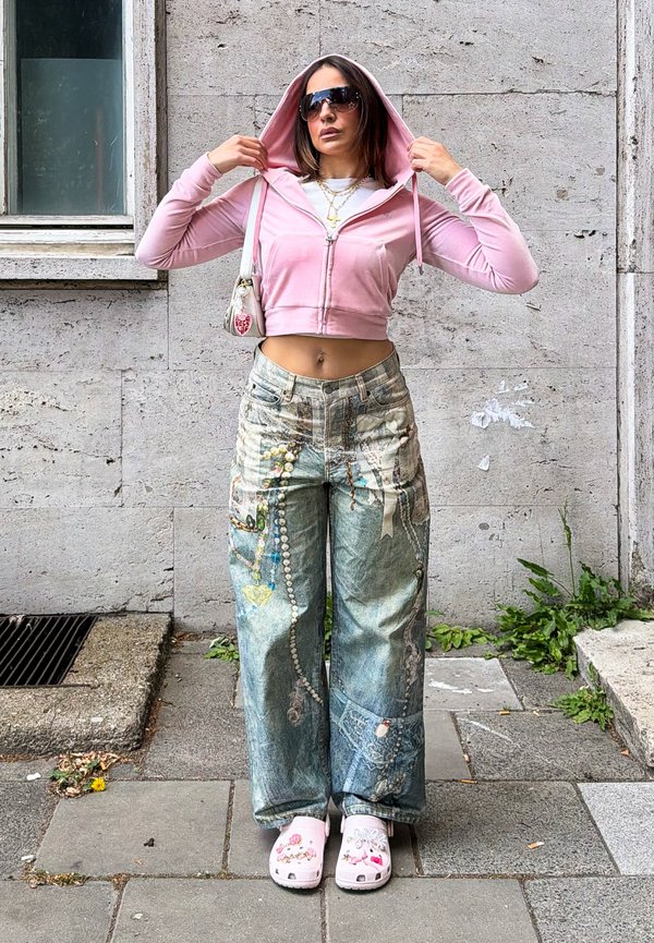 Kvinde iført solbriller, lyserød cropped hættetrøje, brede trykte jeans og lyserøde udsmykkede clogs, stående på en byens fortov.