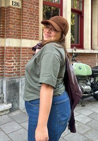 Grøn kortærmet skjorte, blå denim jeans, brun tekstureret kasket og bordeaux læderjakke. Baggrunden har en murstensbygning og en motorcykel.