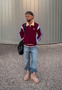 Vínovočervený sveter na zips s pleteným vzorom, béžovým golierom a modrými pruhovanými rukávmi. Svetlomodré rifle a béžové robustné čižmy. Čierna kabelka na rameno.