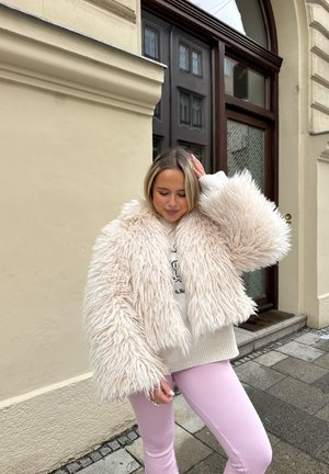 Joven mujer con chaqueta beige esponjosa y pantalones rosas de pie en la acera de la ciudad junto a un edificio con puerta y ventanas de madera.