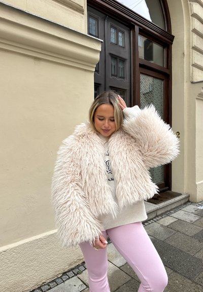 Joven mujer con chaqueta beige esponjosa y pantalones rosas de pie en la acera de la ciudad junto a un edificio con puerta y ventanas de madera.