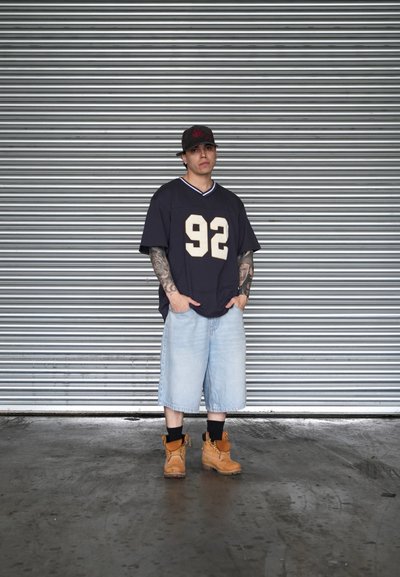 Joven con los brazos tatuados, lleva una gorra negra, una camiseta negra oversize con el número 92, pantalones cortos de mezclilla claros, calcetines negros y botas de trabajo marrones, de pie frente a una persiana metálica.
