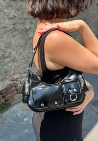 Czarna torebka wykonana z błyszczącej skóry lakierowanej, z dwiema przednimi kieszeniami, srebrnymi akcentami oraz zamknięciem na zamek błyskawiczny. Zawiera regulowany pasek.