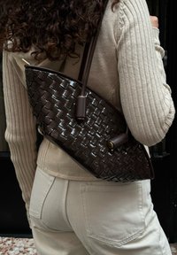Brun vævet lædertaske med en bøjet form, der har glatte detaljer og en lang strap. Båret over skulderen, parret med en lys sweater.