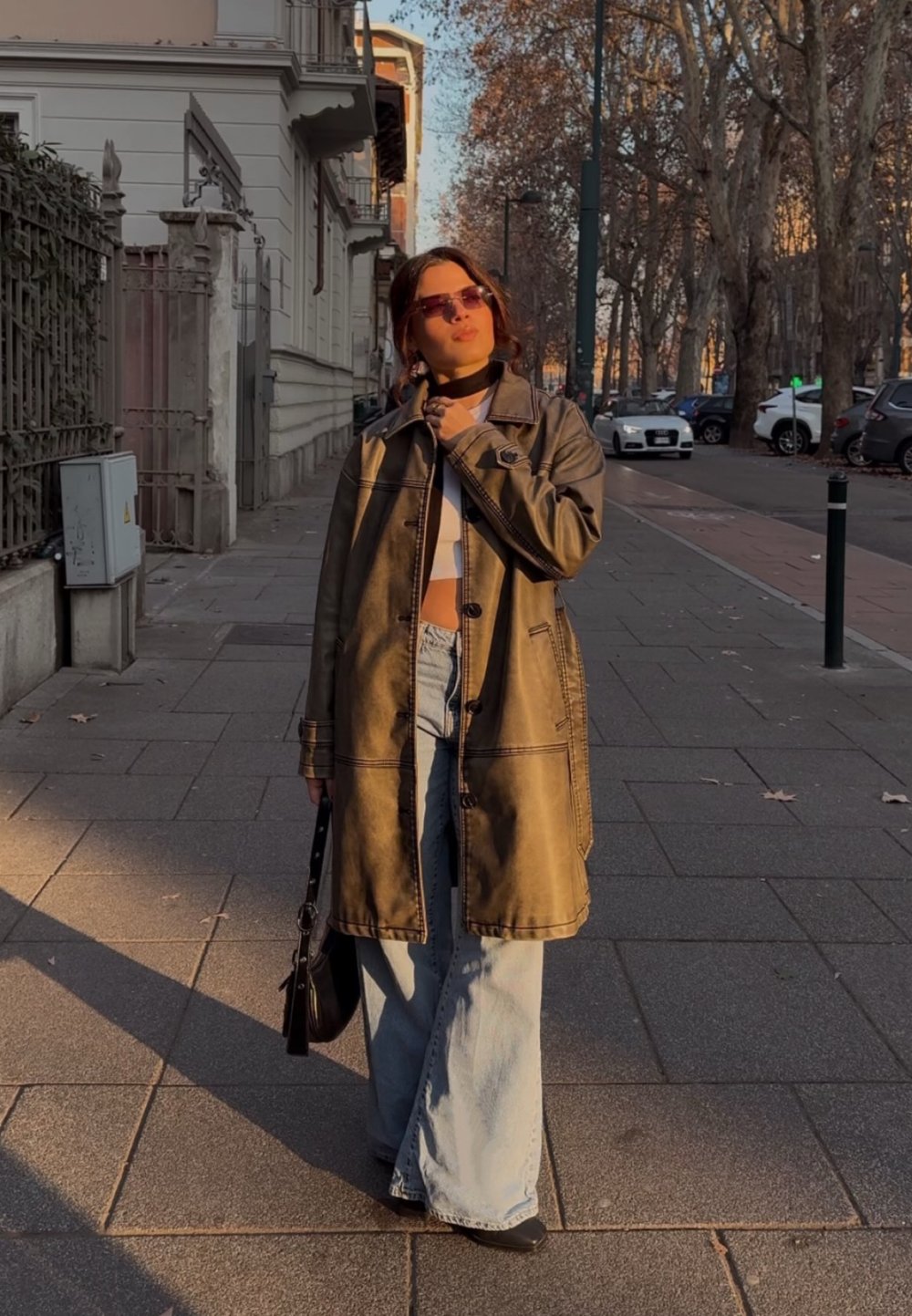 Καφέ δερμάτινο παλτό με γιακά και κουμπιά, φορεμένο πάνω από λευκή μπλούζα και φαρδιά μπλε τζιν παντελόνια. Μαύρη τσάντα χειρός και γυαλιά ηλίου ολοκληρώνουν την εμφάνιση.