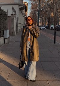 Καφέ δερμάτινο παλτό με γιακά και κουμπιά, φορεμένο πάνω από λευκή μπλούζα και φαρδιά μπλε τζιν παντελόνια. Μαύρη τσάντα χειρός και γυαλιά ηλίου ολοκληρώνουν την εμφάνιση.