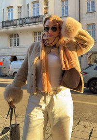 Femme portant un manteau en fausse fourrure beige, une écharpe, des gants, des lunettes de soleil et un pantalon clair, tenant un sac fourre-tout dans une rue ensoleillée avec des bâtiments et des voitures en arrière-plan.