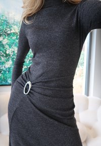 Γυναίκα με εφαρμοστό μακρυμάνικο σκούρο γκρι φόρεμα, με ασημένια οβάλ αγκράφα στη μέση, που στέκεται σε εσωτερικό χώρο κοντά σε παράθυρο και έργα τέχνης.