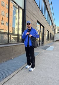 Άτομο που φοράει μπλε αθλητικό μπουφάν με λευκές ρίγες, μαύρο μπλουζάκι, μαύρο παντελόνι και γκρι αθλητικά παπούτσια, κρατώντας μια μαύρη τσάντα έξω από ένα κτήριο.