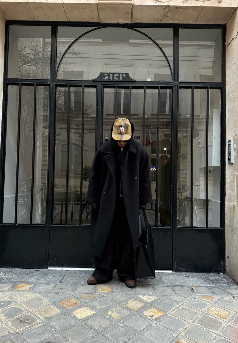 Personne portant un long manteau noir, une casquette à carreaux jaune et des gants, tenant un sac noir, se tenant devant une porte métallique noire avec des panneaux en verre.