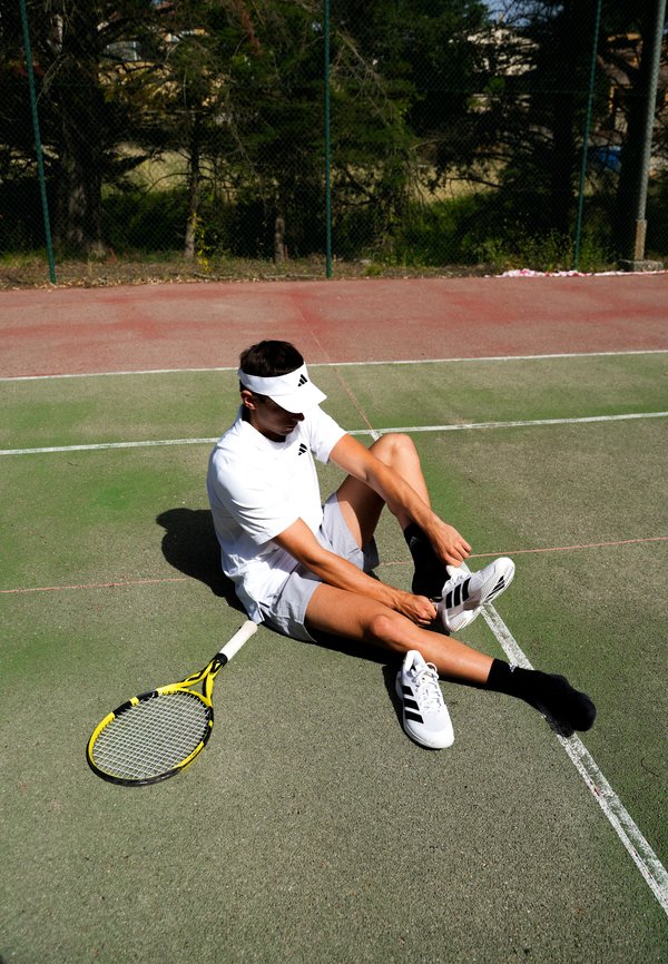 Jeune joueur de tennis masculin assis sur le court en train de mettre sa chaussure, avec une raquette de tennis à côté de lui et portant une tenue de sport blanche et une visière.