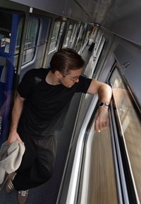 Mees mustas T-särgis ja tumedates pükstes toetub rongiaknale, hoides halli jopet ja kandes randmel kella.