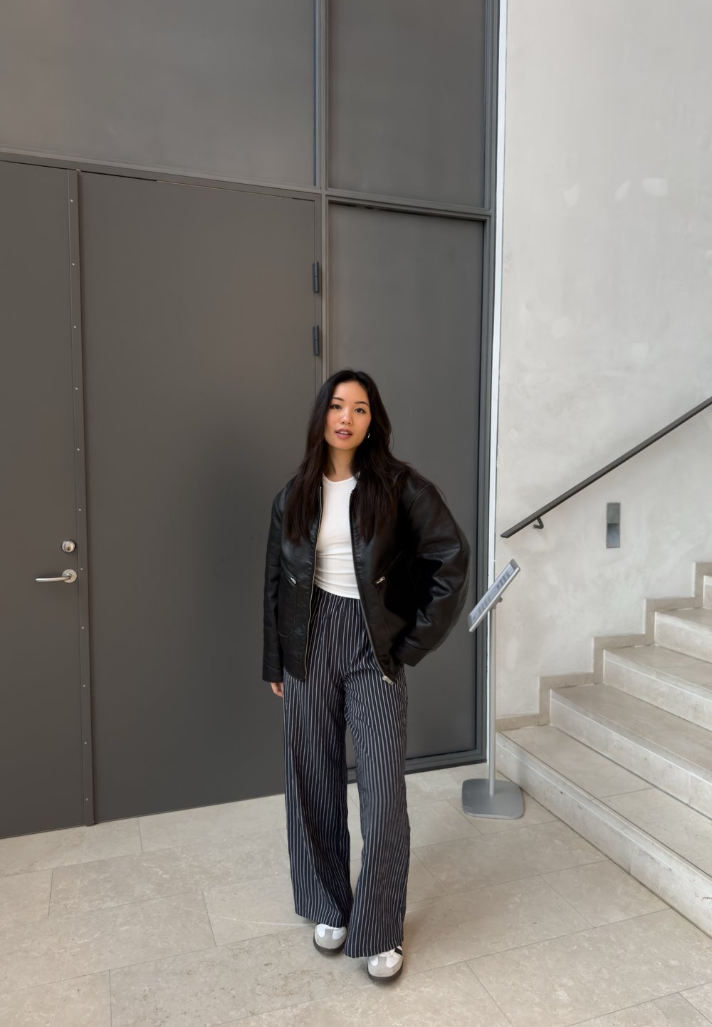 Svart läderjacka, vit skjorta och vida randiga byxor. Grå skor, står nära en grå dörr och en trappa med släta stensteg.