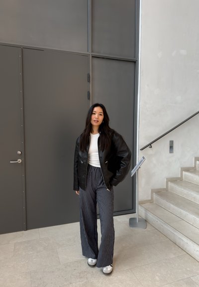 Chaqueta de cuero negra, camisa blanca y pantalones de pierna ancha a rayas. Zapatos grises, de pie cerca de una puerta gris y una escalera con peldaños de piedra lisa.