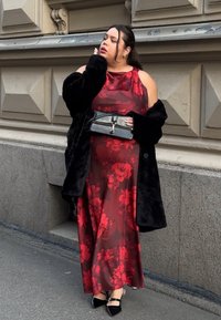 Piros virágos szatén ruha hidegváll kialakítással, fekete műszőrme kabát, fekete táska kapoccsal, és fekete hegyes orrú magassarkú cipő.