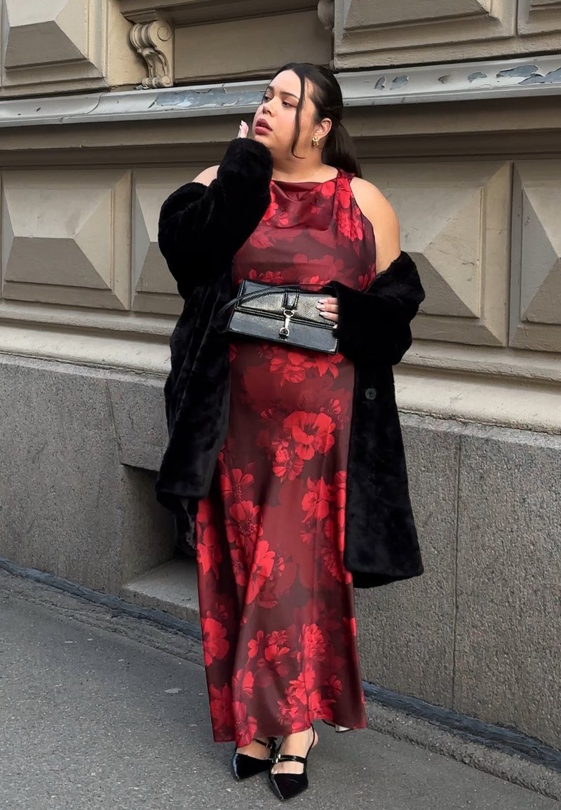 Rød blomstret satengkjole med kald skulder-design, svart kunstpelsjakke, svart håndveske med spenne og svarte spisse hæler.